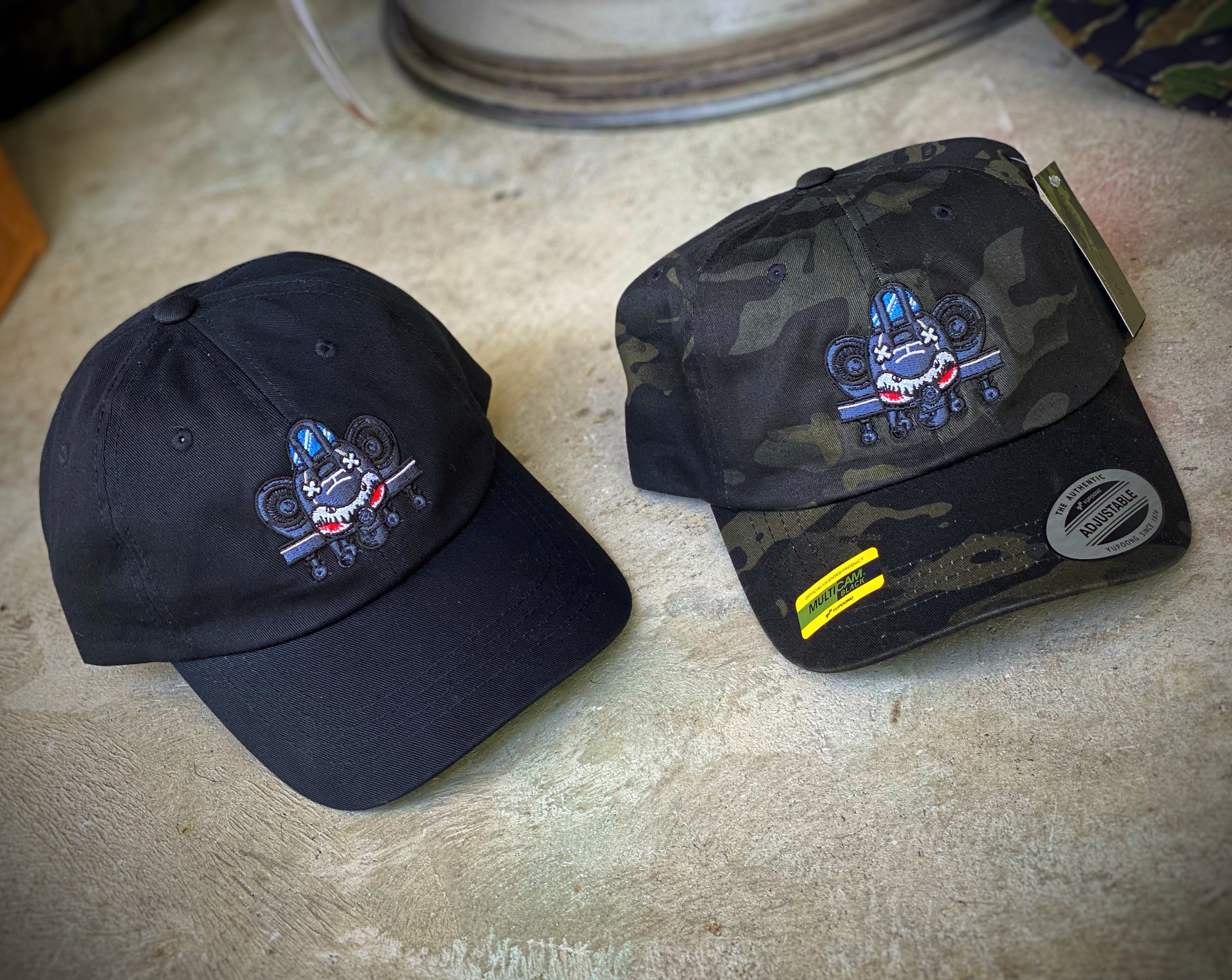One black hat and one Multicam black camo hat both featuring the dangerous goods brand A10 warthog logo embroidered in the center